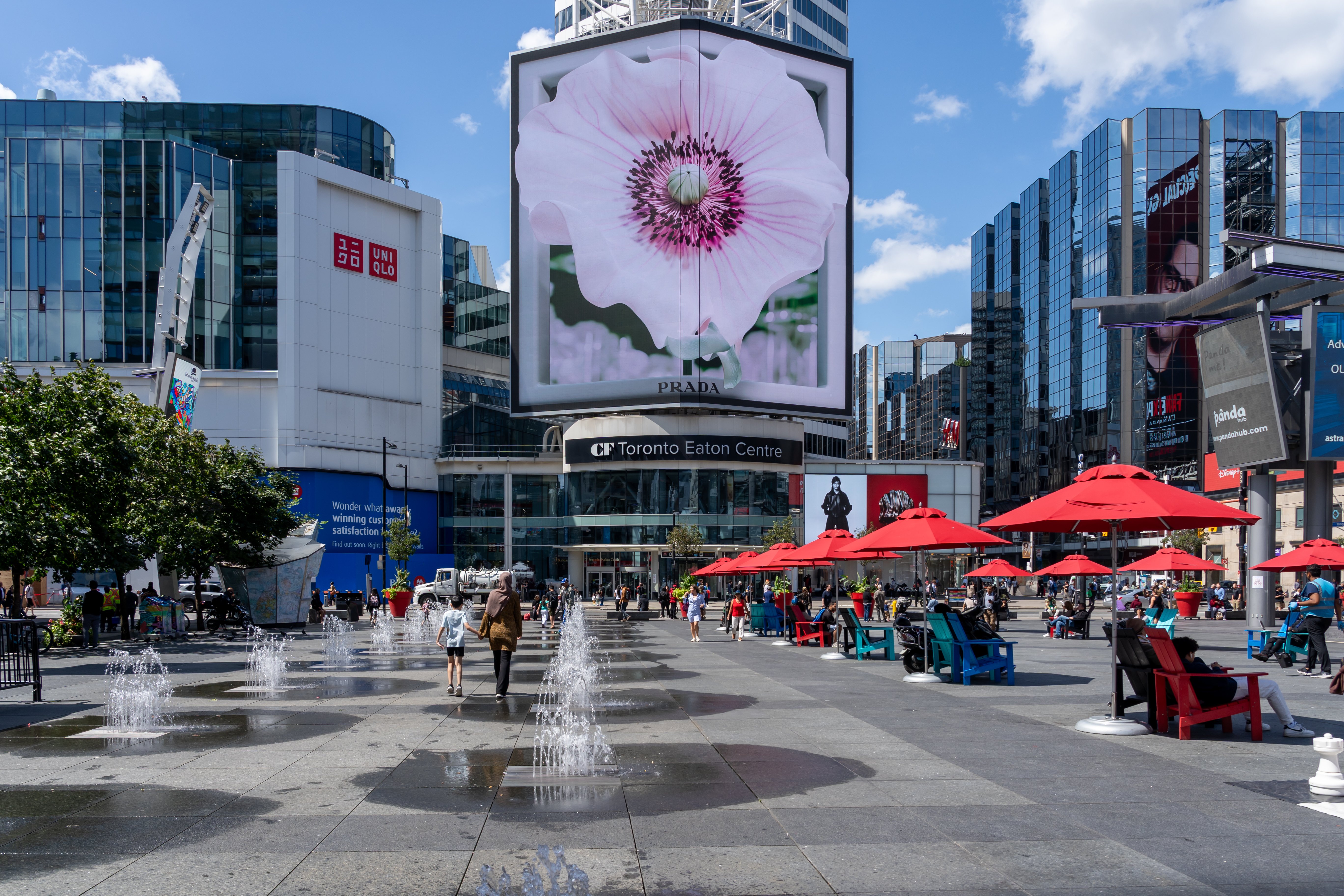 A large digital billboard featuring a Prada advertisement overlooks Toronto’s Yonge-Dundas Square, with pedestrians, fountains and red umbrellas in the foreground.